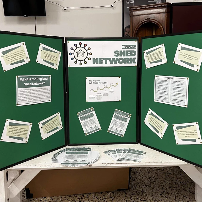 A green three-panel display board promoting the Regional Shed Network at ShedConnect. It includes information about the network, its aims and feedback from participants, with leaflets arranged on the table below. The display highlights support, connection and opportunities for collaboration between sheds.
