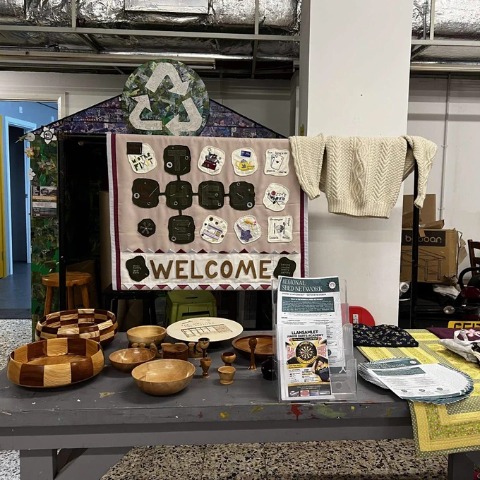A display table at ShedConnect featuring handmade wooden bowls and small turned items, alongside a knitted cream jumper, fabric items and leaflets. Behind the table is a “Welcome” board with sewn and quilted pieces, highlighting creativity and crafts produced by Men’s Sheds members.
