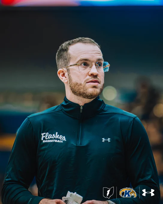 Kent State men's basketball Assistant Coach Jon Fleming, wearing glasses and a black "Flashes Basketball" Under Armour quarter-zip, looks intently toward the court before a game.