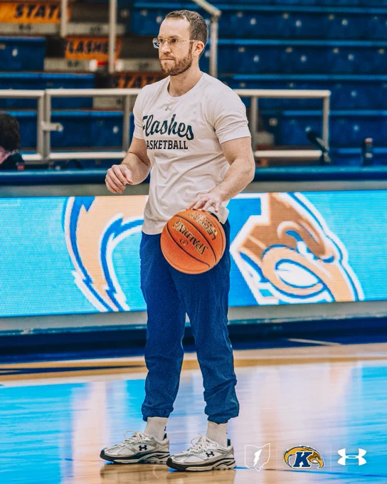 Kent State men's basketball Assistant Coach Jon Fleming, wearing glasses and a white "Flashes Basketball" Under Armour t-shirt with blue sweatpants, holds a Spalding basketball on the court during pregame warmups, with the Kent State arena LED boards visible in the background.