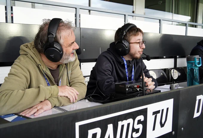 Two audio descriptive commentators at Pride Park Stadium wearing headsets and microphones, seated on the press bench.