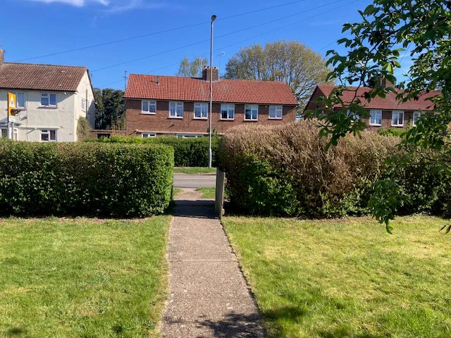 Front lawn with freshly cut green grass, bordered by neatly trimmed hedges, with houses and a blue sky in the background.