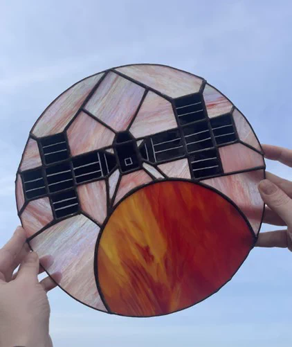 A circular stained-glass panel shows the Psyche spacecraft passing close to Mars. The planet glows in layered reds, oranges, and golds, while the spacecraft appears in lighter metallic tones against dark space. The warm colors surround the craft briefly, suggesting a close flyby before it continues into the darkness beyond.