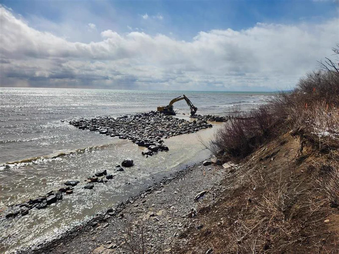 An excavator works on a rocky shoreline of a lake, placing large stones into the water to create a protective groyne against erosion under a cloudy sky.