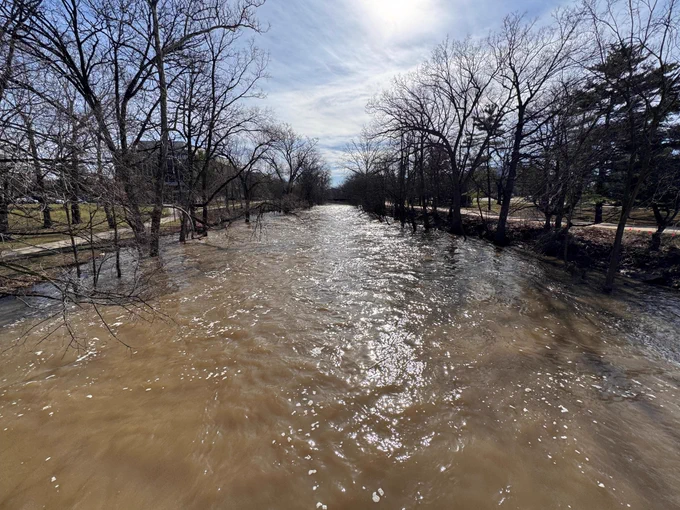 A wide muddy river flows between bare trees under a sky with light clouds. The water reflects the sky and trees.