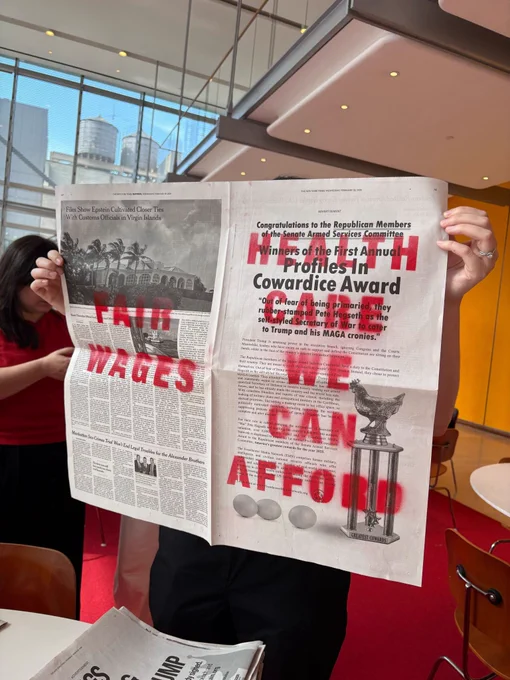 A print copy of The New York Times, spray painted in red. On one side it says "FAIR WAGES." On the other, it says "HEALTH CARE WE CAN AFFORD"