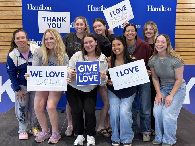 A group of people smiling and holding up signs with messages like "Thank You," "I Love Hamilton," "We Love Softball," and "Give to What You Love." They are standing in front of a blue backdrop with "Hamilton" written multiple times.
