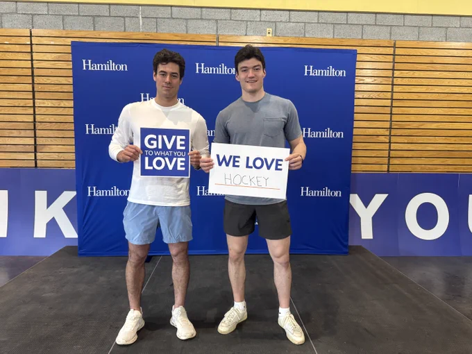 Two individuals standing in front of a backdrop with "Hamilton" written on it. One person holds a sign that reads "Give to What You Love," and the other holds a sign saying "We Love Hockey.