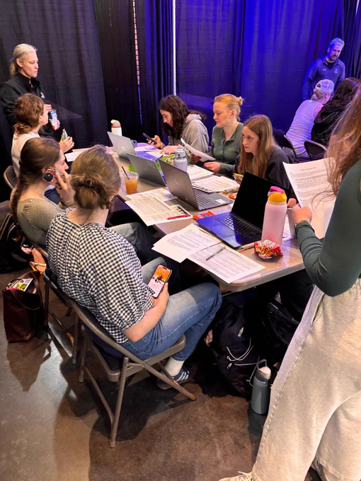 A group of people sit around a table equipped with laptops and documents, some using their phones. Snacks and drinks are scattered across the table. A curtain and blue lighting are visible in the background.