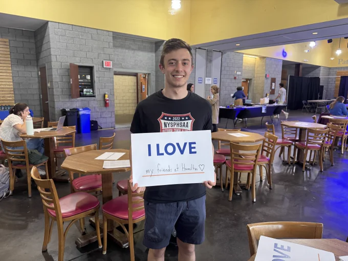 A person stands in a cafeteria holding a sign that reads, "I love my friends at Hamilton" with a heart symbol. The surroundings include wooden tables and chairs, and there are other people in the background engaged in various activities.