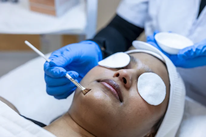 A person is lying on a bed and has coverings over their eyes as another person uses a brush on their skin.
