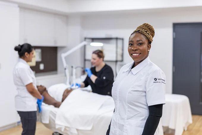 A person stands in front of two other people providing treatment to a client who’s lying on a bed.
