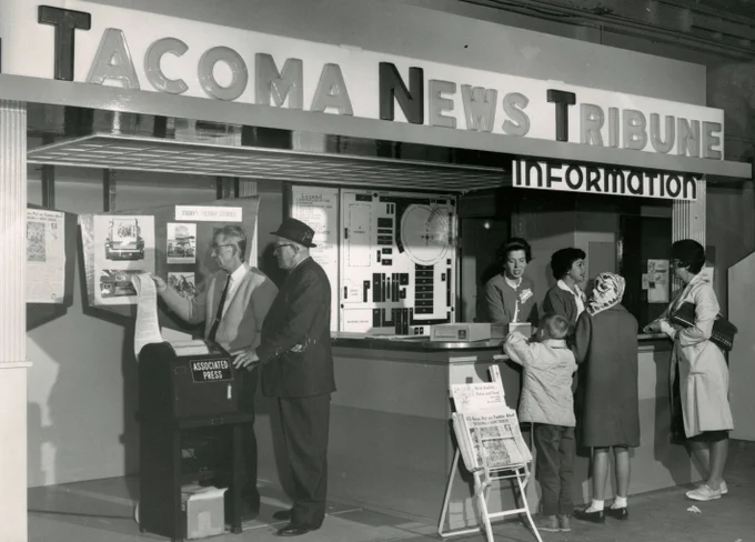 Black-and-white photo of a Tacoma News Tribune exhibit at the Western Washington Fair, 1964. A large overhead sign reads “Tacoma News Tribune,” with an “Information” counter beneath it. At left, two men—one in glasses and a cardigan, the other in a hat and overcoat—stand beside an Associated Press machine displaying photos and news pages. At right, three women and a young boy gather at the counter, speaking with a staff member. A rack of newspapers and display boards with photos and diagrams are visible in the background.
(Courtesy Tacoma News Tribune, Image TNT0122N, Tacoma Public Library Northwest Room)