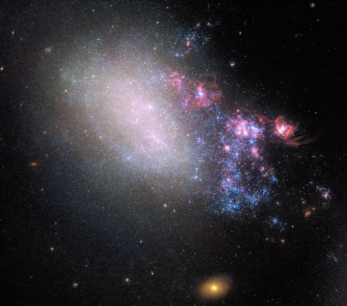 A hazy galaxy shines against black space, with brighter blue stars seen at right, interspersed with patchy clouds of pink nebulae.