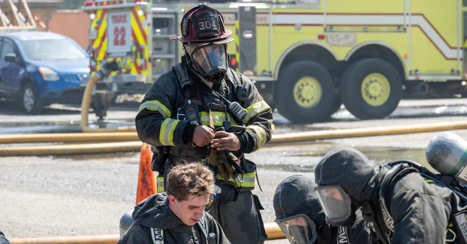 Firefighter in full gear adjusting equipment near tthree crouching colleagues with fire truck in background.
