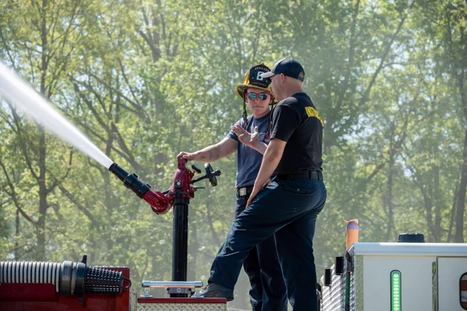 Two firefighters operate a fire hose nozzle spraying water outdoors with trees in the background.