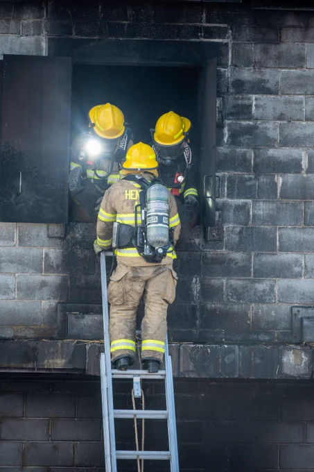 Three firefighters in full gear and yellow helmets by a dark window of a charred brick building using a ladder during a rescue operation.