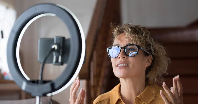 Person recording a video using a smartphone mounted on a ring light stand, wearing glasses and smiling, indoors.