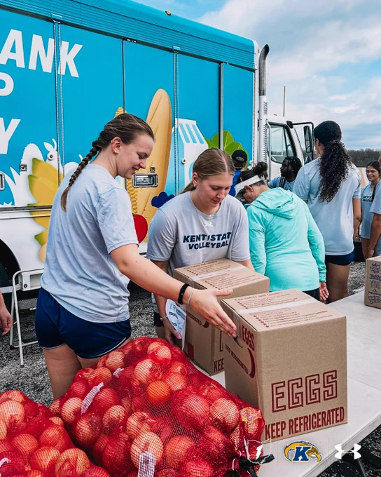 A group of individuals is volunteering near a colorful truck labeled with a food bank sign. They are organizing bags of apples and boxes labeled "Eggs Keep Refrigerated." One of the individuals is wearing a "Kent State Volleyball" shirt. Two logos, possibly representing sponsors, are visible at the bottom, including the Under Armour logo.