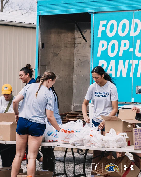 Four individuals are organizing food items in front of a blue "Food Pop-Up Pantry" truck. They are sorting groceries into bags and boxes. The side of the truck prominently features the Kent State University logo.