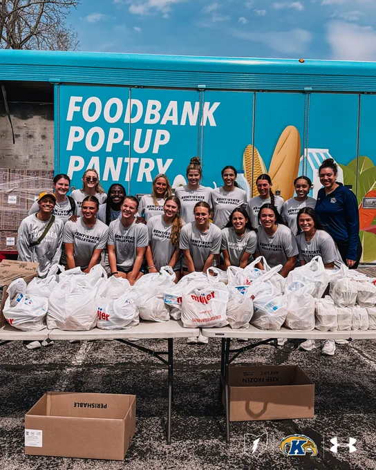 A group of people in matching team shirts stand in front of a "Foodbank Pop-Up Pantry" truck. They are smiling and pose behind a table filled with grocery bags. The image includes the logos of Kent State University and Under Armour.