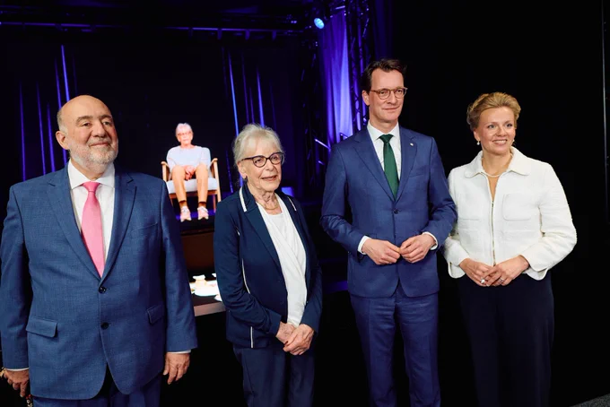 Ministerpräsident Hendrik Wüst, Ministerin Ina Brandes, ein älterer Mann und eine ältere Frau stehen nebeneinander vor einer Bühne mit dunklem Hintergrund und violetter Beleuchtung. Im Hintergrund ist ein Hologramm einer älteren Frau zu sehen, die auf einem Stuhl sitzt.
