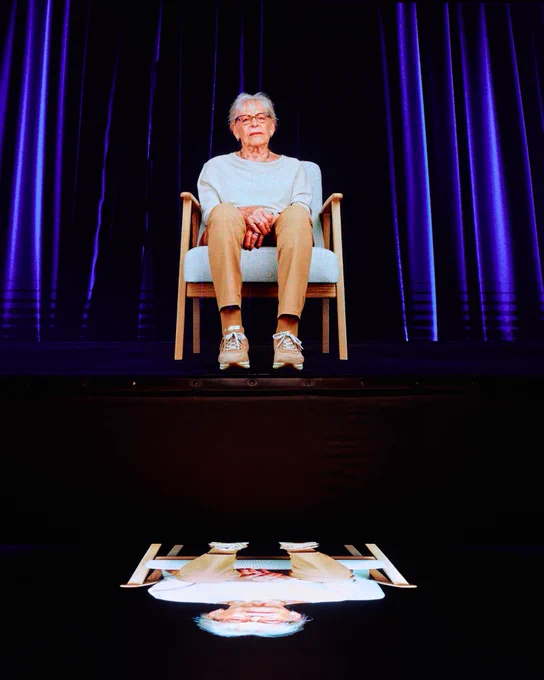 Ein Hologramm auf einer Bühne vor violett beleuchteten Vorhängen zeigt eine ältere Frau, die auf einem Stuhl sitzt.