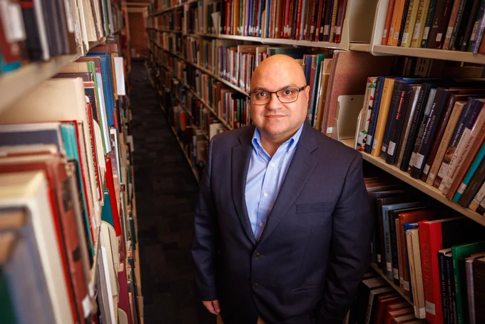 Bedross Der Matossian, Hymen Rosen Professor in Judaic Studies at the University of Nebraska–Lincoln, stands between rows of bookshelves in Love Library. He is wearing a dark blue sport coat over a light blue dress shirt, and glasses. (Jordan Opp/University Communication and Marketing)