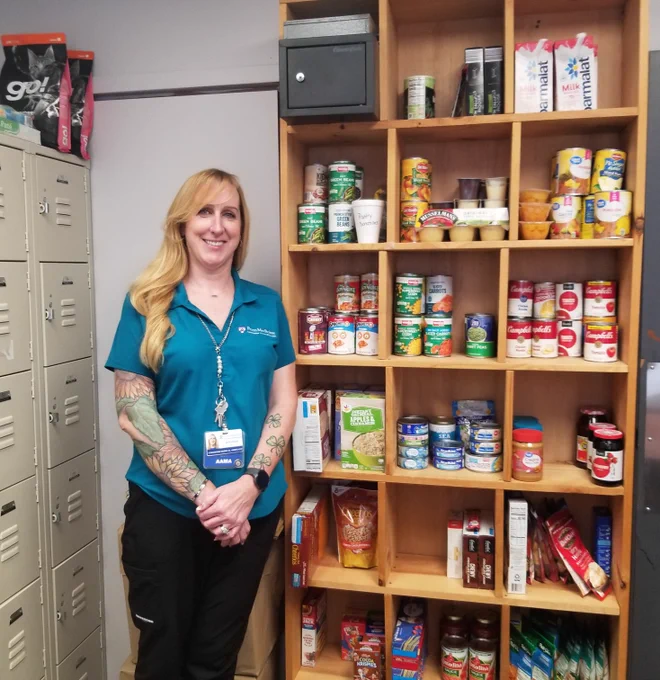 A woman stands near a wooden shelf stocked with food items like canned goods, cereal boxes, and Parmalat milk, along with pet food. She wears a blue scrub top, black pants, and a badge labeled \"AAMA.\"