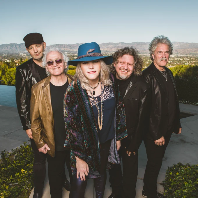 Five members of the Jefferson Starship Band stand outdoors, smiling. They are dressed in casual, stylish outfits with a scenic landscape in the background.