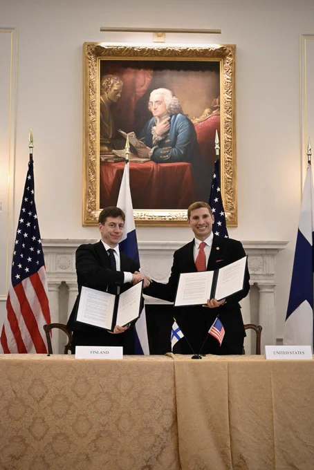 Minister and State Dept representative Helberg hold up signed documents and shake hands in front of a large painting, a fireplace, and US and Finnish flags.
