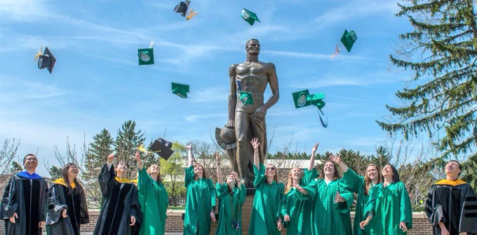 Image of Michigan State University graduates dressed in graduation regalia, throwing caps in the air outside of the Spartan Statue.