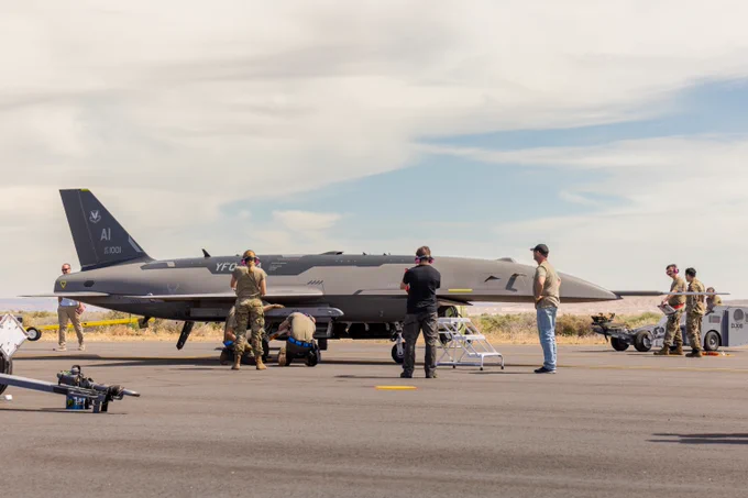 Military personnel inspect a sleek, gray unmanned aerial vehicle on a runway under a partly cloudy sky.