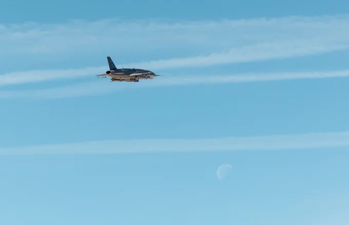 Military jet flying through a clear blue sky with faint contrails and a visible moon in the background.