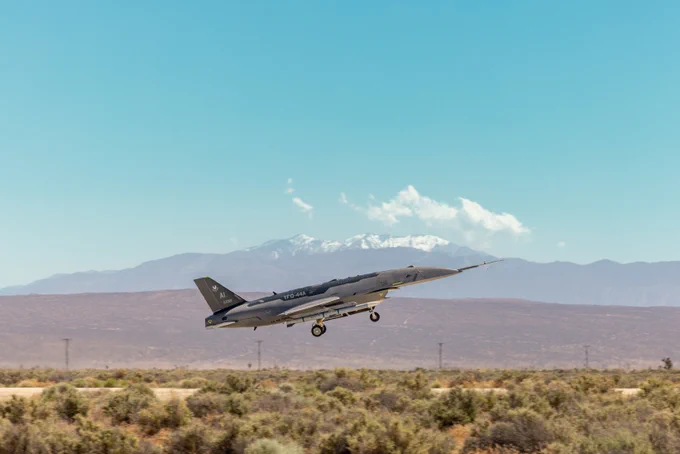 Military jet taking off from desert runway with mountains in the background under clear blue sky.
