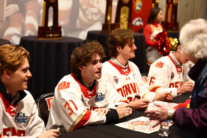 Three individuals wearing Denver hockey jerseys are seated at a table signing autographs. Championship trophies are displayed in the background. Fans are interacting with the individuals.