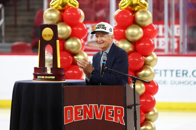 A person standing at a podium labeled "DENVER" is clapping. Nearby, there is a large NCAA National Champion trophy. Behind the podium, there are red and gold balloon arrangements. The event seems to be taking place on an ice rink.