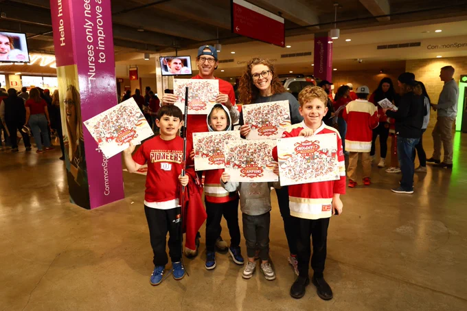 A group of people, including both adults and children, is standing inside an entertainment venue. They are holding posters featuring the DU Hockey team. In the background, other people are mingling, and there are overhead signs and kiosk areas visible.