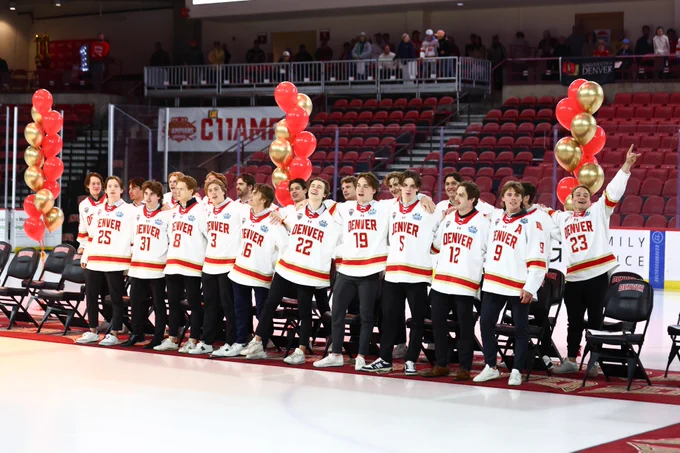 A group of hockey players wearing jerseys with "Denver" stand together on an ice rink. The scene includes red and gold balloons. The stands in the background are mostly empty, and chairs are arranged on the ice in front of the players.