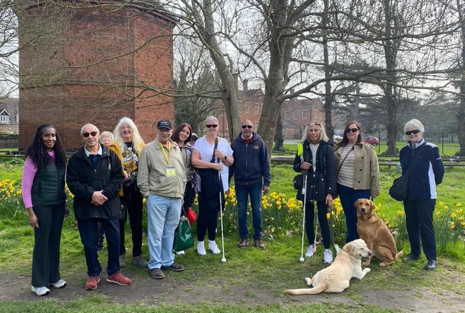 A group of visually impaired adults lined up smiling on a sunny day with  two people holding white canes and two guide dogs sitting and laying on the grass in front.