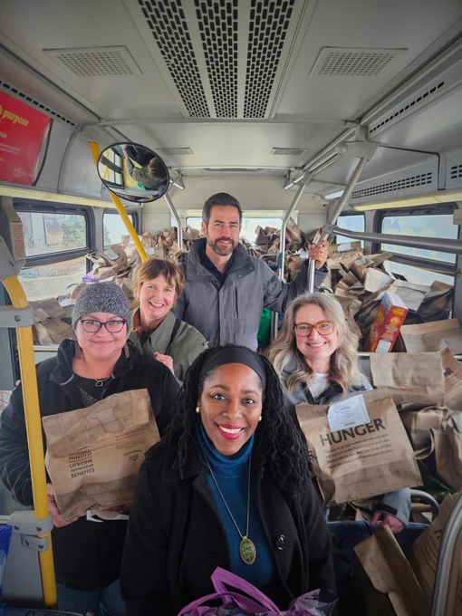 Five members of the Co-operatives First team - four women and one man - hold bags of donated food inside a city bus for the Stuff the Bus campaign.