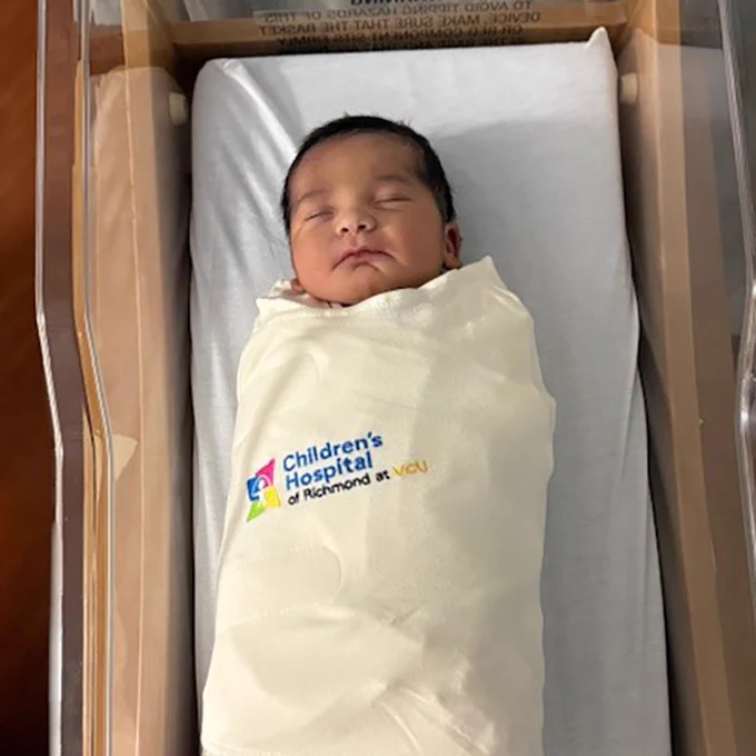 A newborn wrapped in a light-colored blanket, peacefully asleep in a hospital bassinet. The blanket displays the logo of the Children's Hospital of Richmond at VCU.