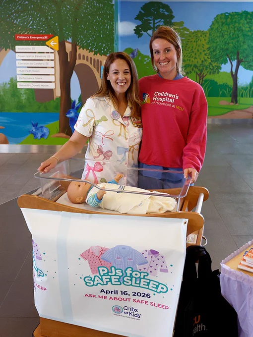 Two individuals stand beside a hospital crib with a baby doll inside. They are in front of a colorful mural at Children’s Hospital of Richmond at VCU. The crib has a "PJs for Safe Sleep" banner, promoting an event on April 16, 2026.