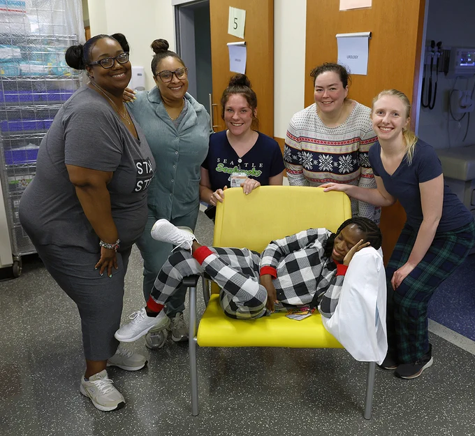 A group of six people smiling, with one person sitting on a yellow chair, dressed in black and white checkered pajamas. The others are standing around the chair in casual attire, including festive pajamas. The setting appears to be a medical setting.