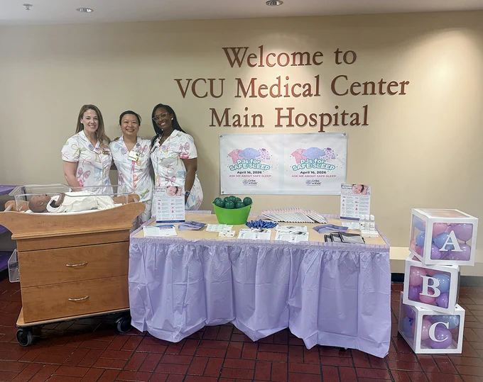 A group of three people stands beside a table display at VCU Medical Center Main Hospital. The table features decorative blocks labeled with "A," "B," and "C." Informational materials and a container of green items are on the table. A baby mannequin is in a crib on the left. Posters on the wall behind them say "Back to Sleep, Every Sleep" with event details.