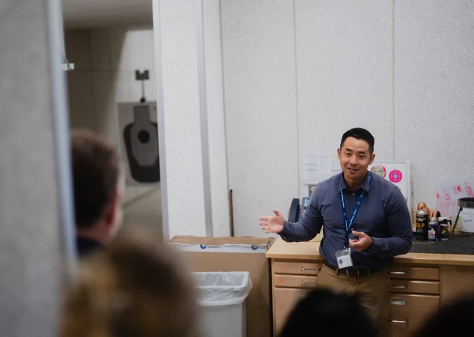 Kern Regional Crime Lab Director Garett Sugimoto gives a presentation in a room with firearms training targets visible in the background. A counter with pamphlets and signs is beside them. An audience listens attentively in the foreground.