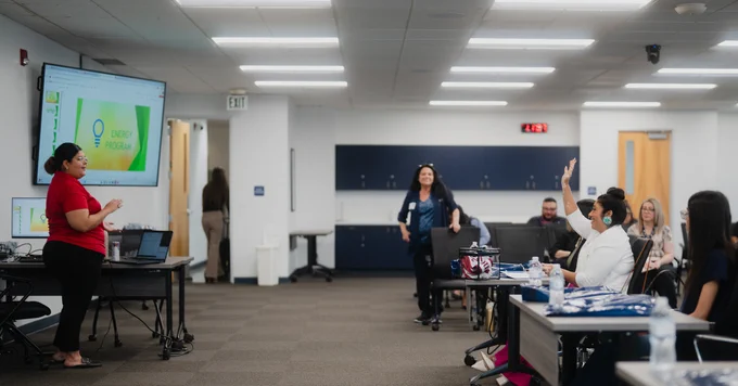 CAPK's Savanahh Oates stands near a large screen displaying a presentation titled "Energy Program" in CAPK's board room. Several people, both CAPK and RAMP folks, are seated at desks, with one person raising their hand. A person stands facing the group.