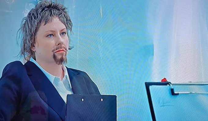 Photo of comedian Amy Gledhill dressed as Alex Horne from the TV show Taskmaster. She is wearing a dark navy blazer over a white collared shirt and holds a black clipboard. She features a prosthetic brown goatee and mustache and has short, messy ash-brown hair. She maintains a serious, deadpan expression while looking off-camera against a bright, blurred blue and white background.