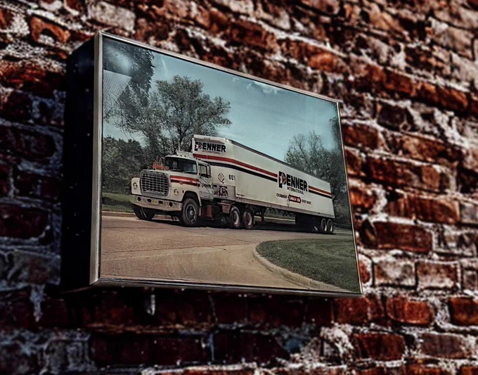 A framed vintage photograph is mounted on a textured red brick wall. The photo shows a classic Penner International transport truck with a white trailer and red Penner logo parked on a curved road. Trees and greenery fill the background, giving the image a nostalgic and historical feel.