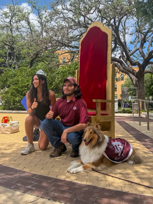 Two people are crouched next to Reveille X, who is wearing a maroon outfit with the university's logo. They are giving thumbs-up gestures. Behind them is a large ornamental throne. Trees and a building are in the background.